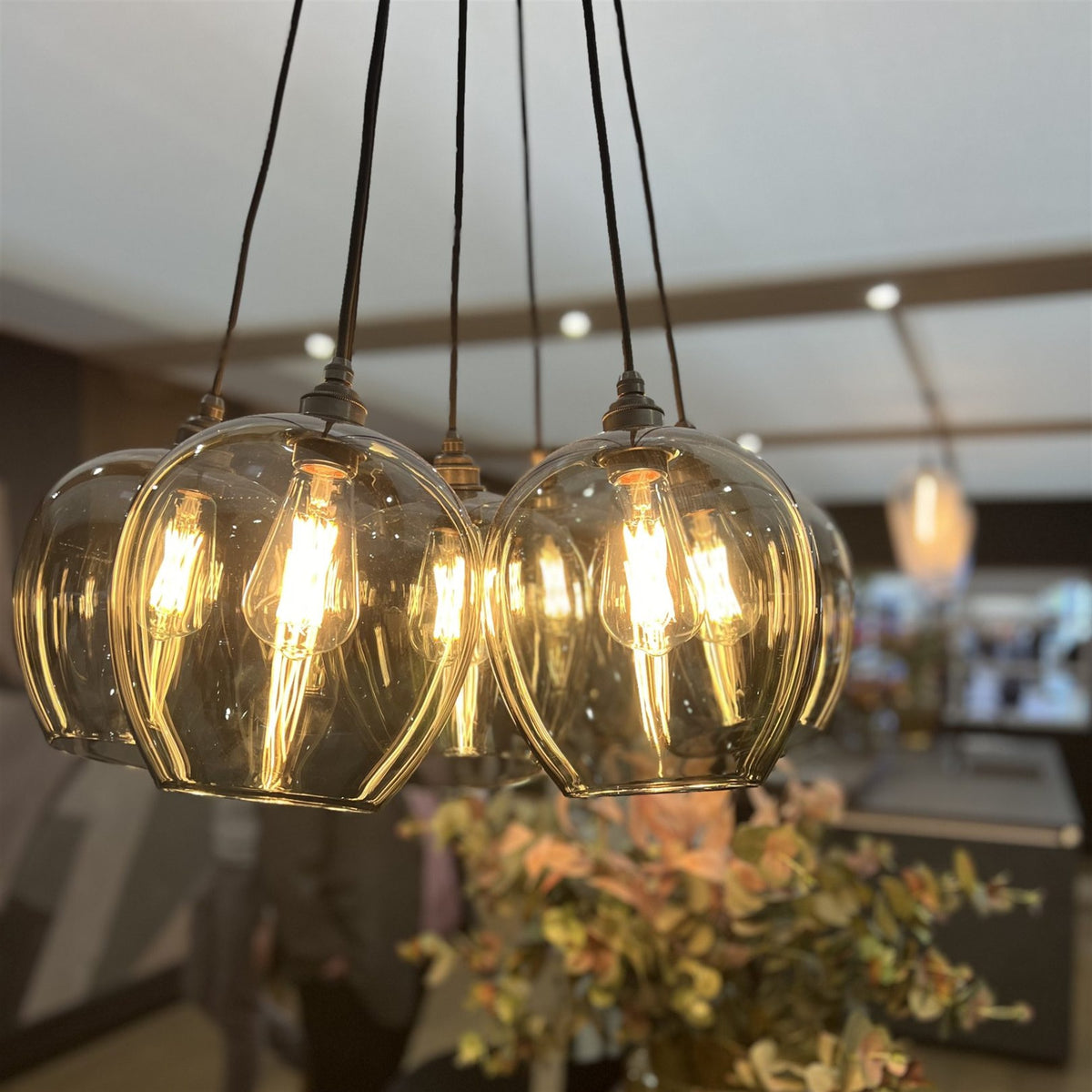 Cluster of tinted clear glass pendants with lit filament bulbs hanging together on dark flex, with a blurred room behind