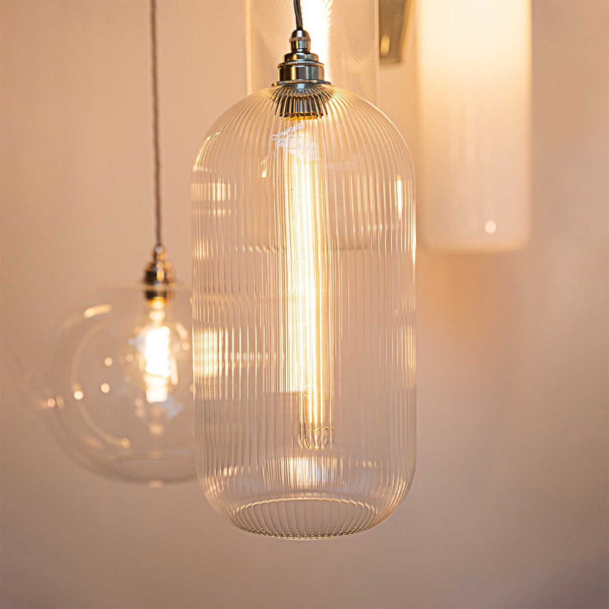 Close up of a lit Leverint Charlton pendant in ribbed glass with a nickel toned fitting, a round glass pendant and a wall light softly blurred behind