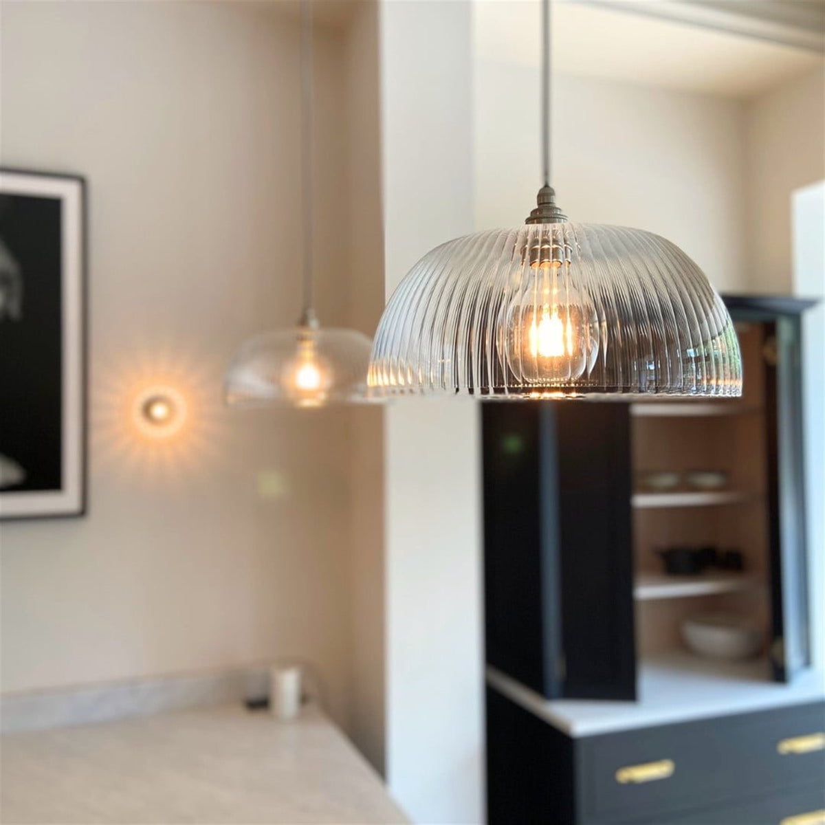 Ribbed glass dome pendant lit with a filament bulb in a kitchen beside a framed picture, with a second clear glass pendant out of focus behind