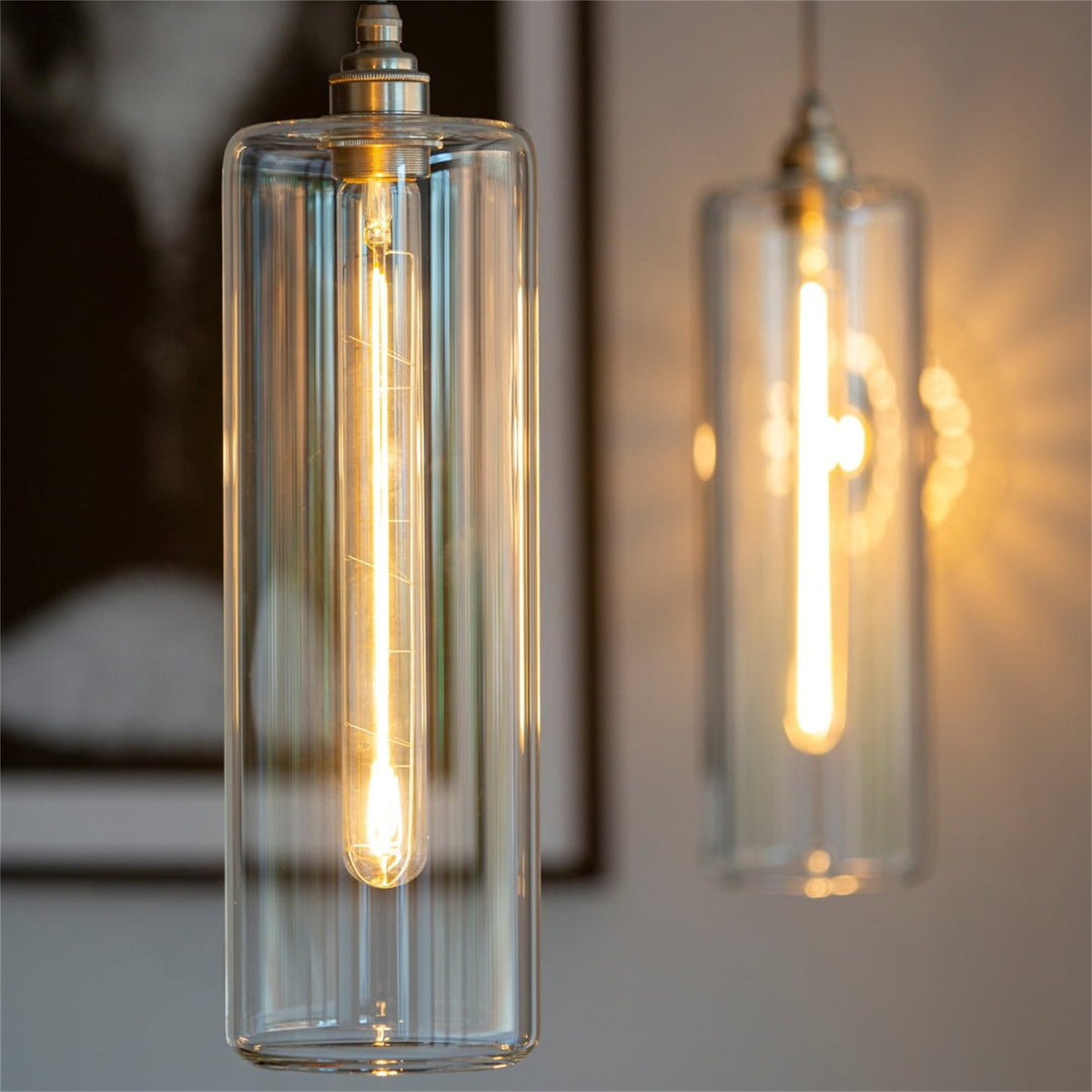 Close up of two tall clear glass cylinder pendants from a Leverint Piccadilly cascade cluster, each with a long filament bulb lit inside, silver toned lamp holder at the top