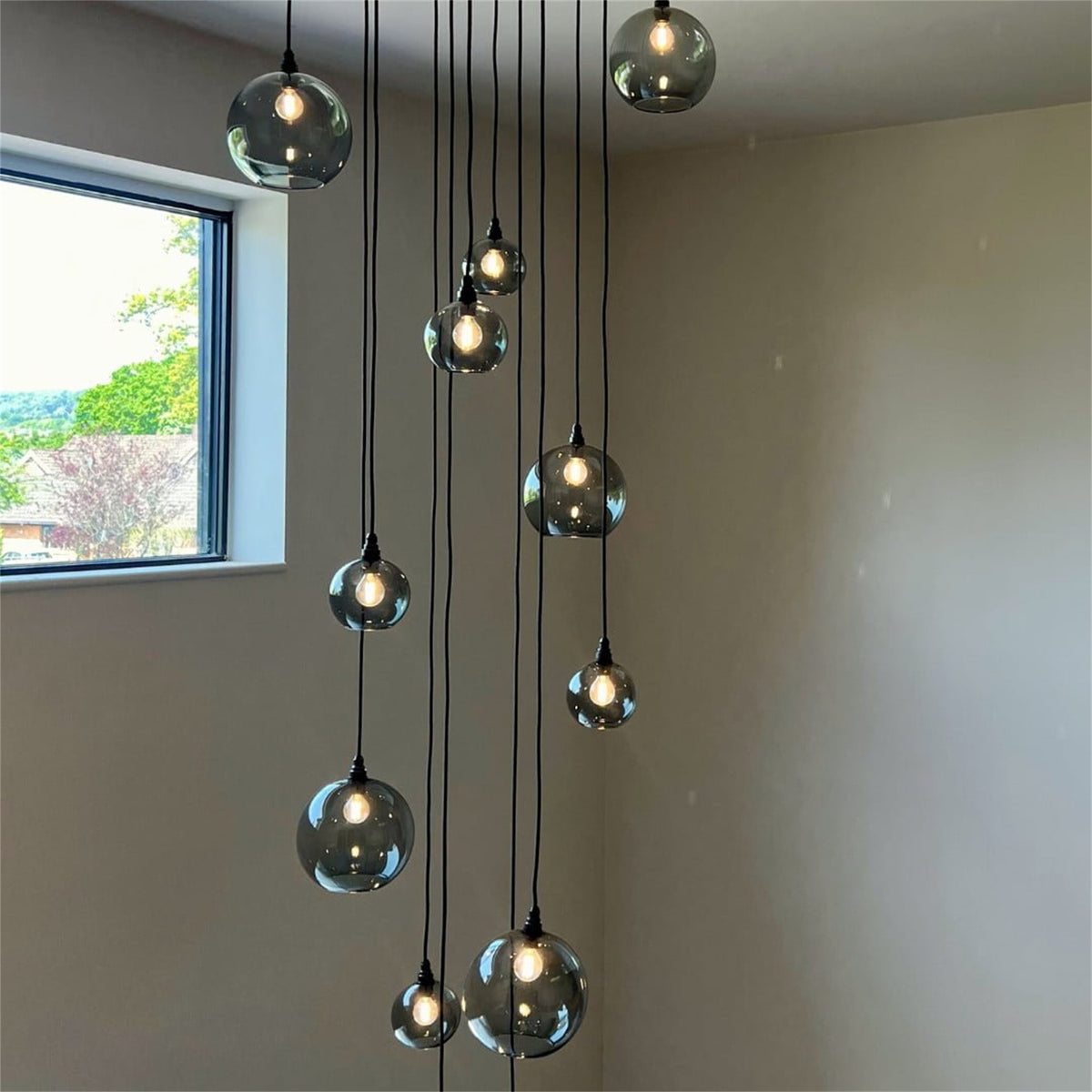 Leverint Pimlico cascade cluster of smoky grey glass globe pendants hung at varied heights on black cables in a tall room beside a window, bulbs lit