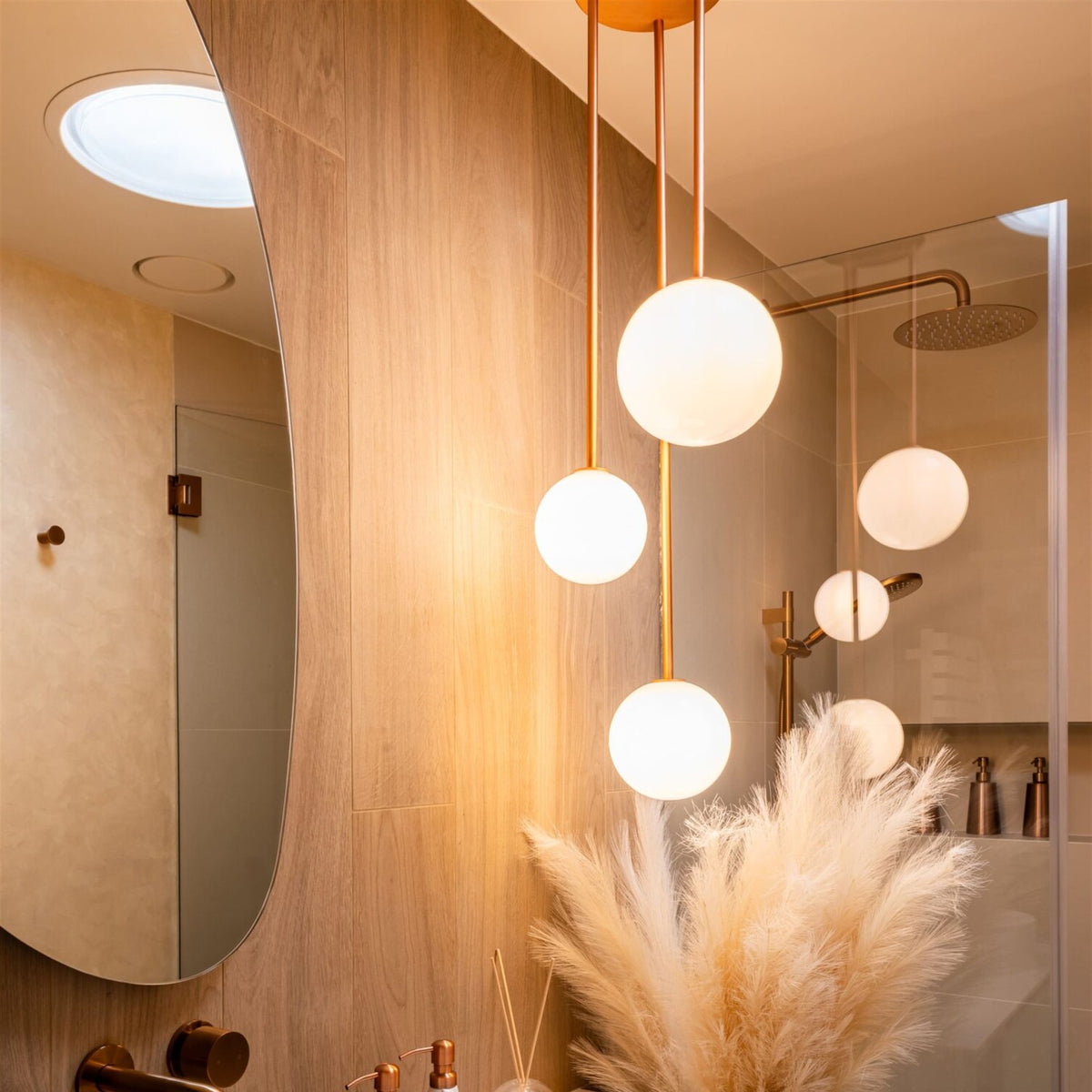 Three opal white glass globes on copper coloured rods glowing above a ribbed white basin and green vanity unit, with pampas grass beside them