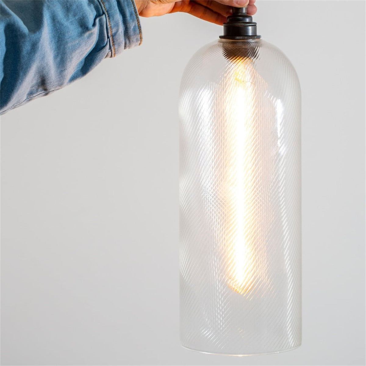 Hand holding a lit Leverint Richmond pendant in clear glass with fine diagonal ribs and a black fitting, against a plain grey wall