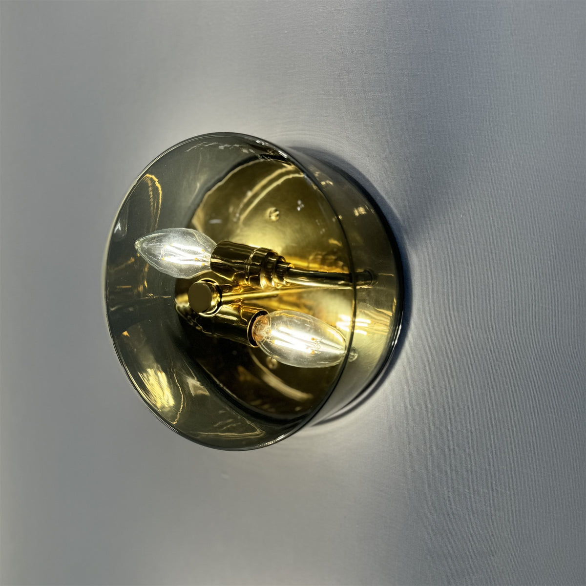 Leverint Temple Flush Mount in smoky transparent black glass with a brass interior and two lit candle bulbs, against a pale grey wall