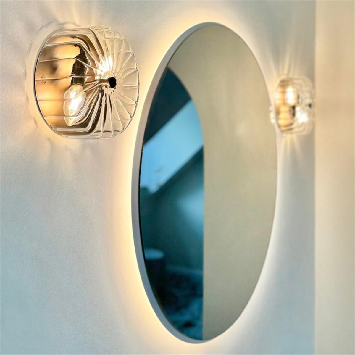 Two Leverint Temple Flush Mounts in clear ribbed glass lit on a cream wall either side of an oval backlit mirror