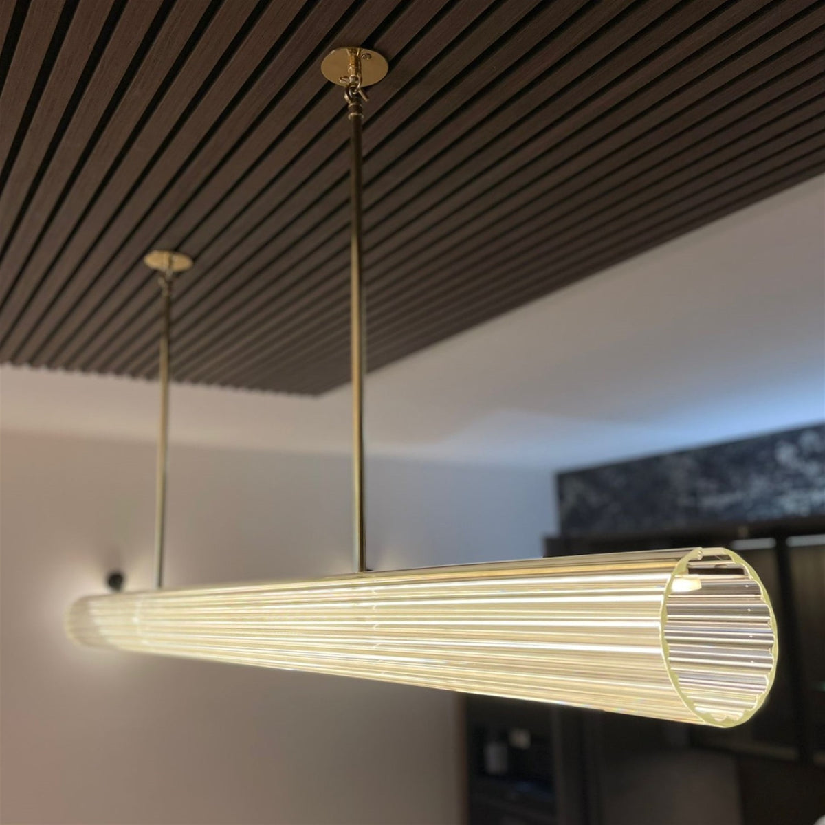 Close up of the end of a Leverint Temple Rod Pendant, a ribbed glass bar lit warm and hung from slim brass rods against a slatted timber ceiling (lifestyle photo, length may differ from the XL)