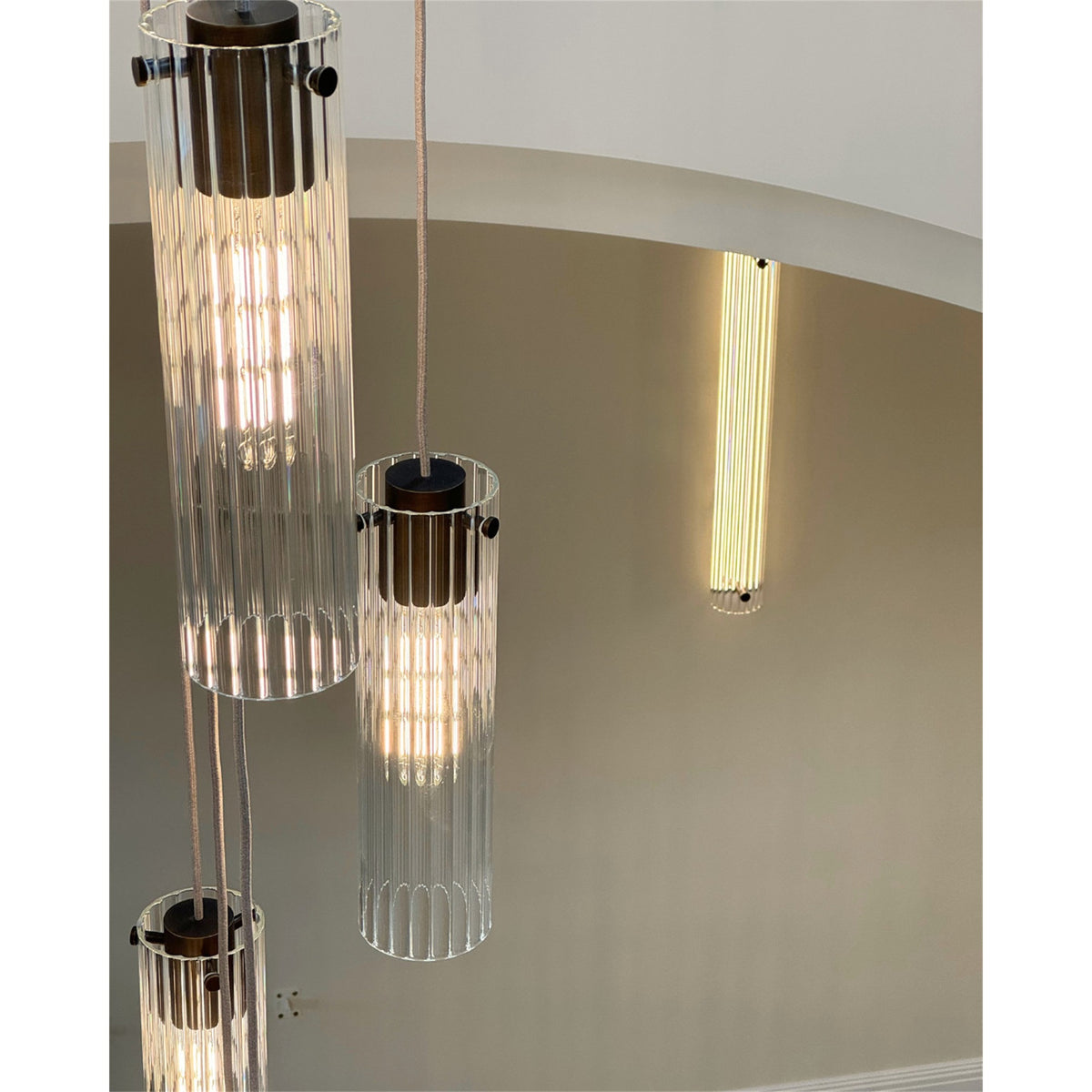 Close up of three fluted glass cylinder pendants with dark bronze tops on fabric covered flex, with a lit fluted glass wall light in the background