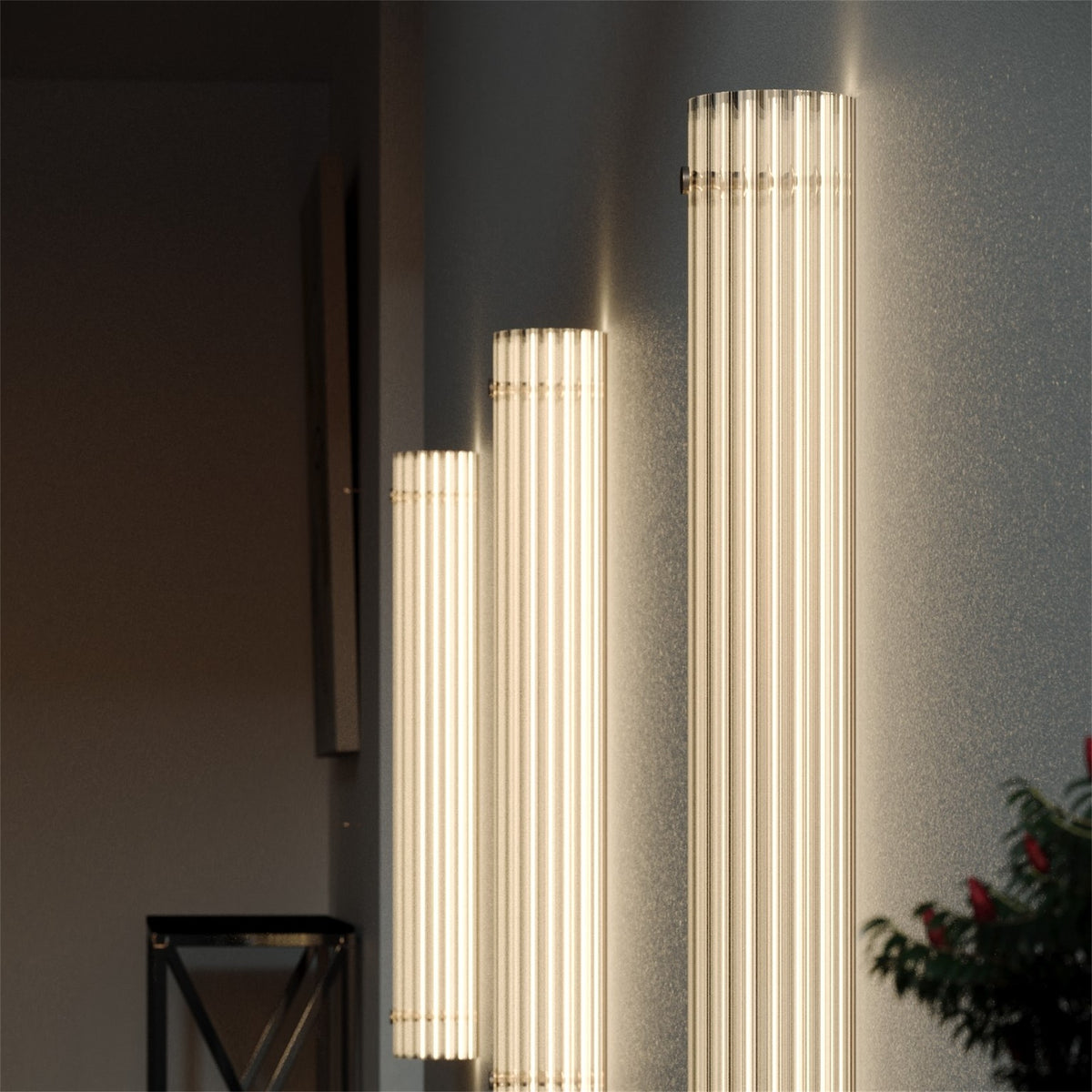 Three lit Leverint Temple Pillar Lights stepped in height on a dark grey wall, ribbed glass columns glowing, with a plant in the corner