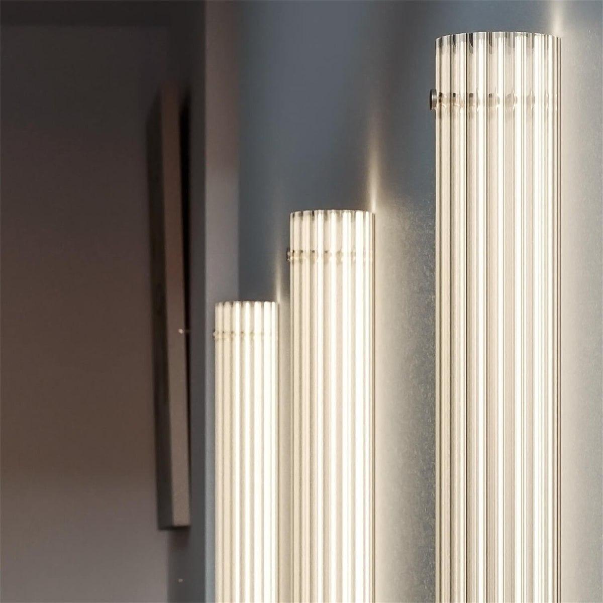 Three lit Leverint Temple Pillar Lights of different heights on a dark grey wall, close up of the glowing ribbed glass columns