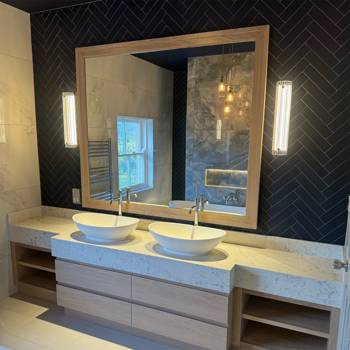 Pair of lit Leverint Temple Pillar Lights either side of a large wood framed mirror above a double vanity, on dark herringbone bathroom tiles