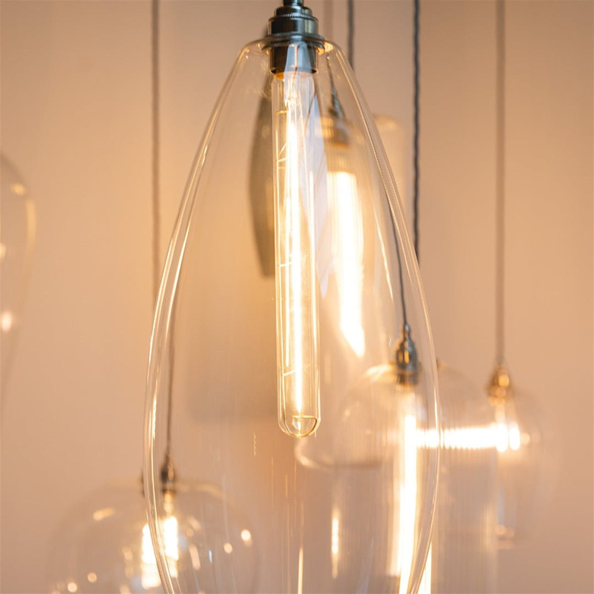 Close up of a slim oval clear glass pendant with a long tubular filament bulb and a polished metal lamp holder, other glass shades softly blurred behind
