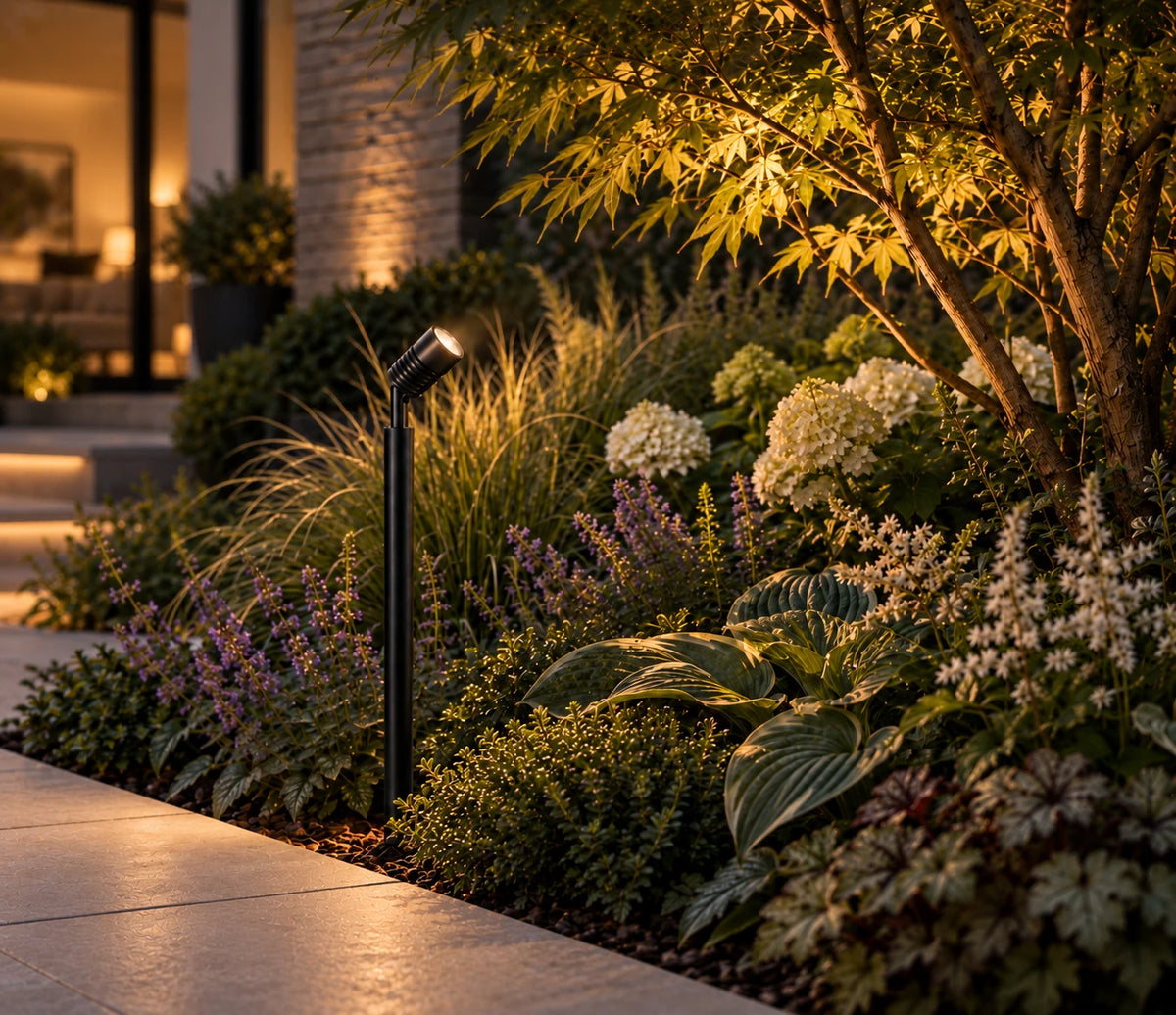 LuxR black powder coated pole spot in a planted garden border at dusk, beside a paved path and a lit maple tree