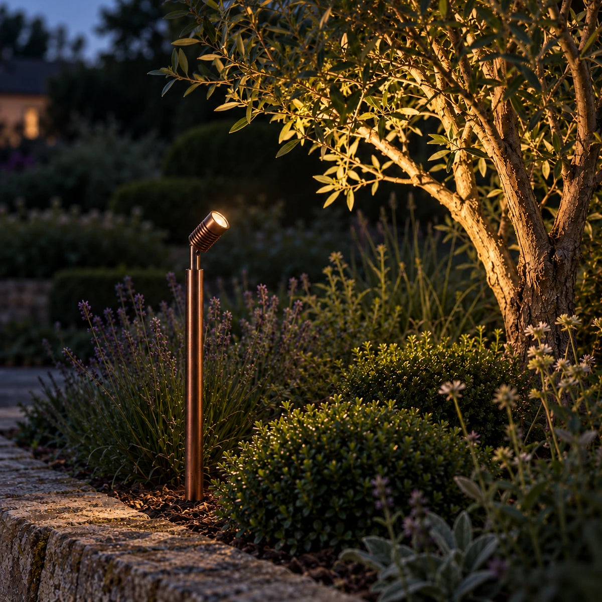 LuxR copper pole spot in a planted garden border at dusk, lighting the branches of a tree