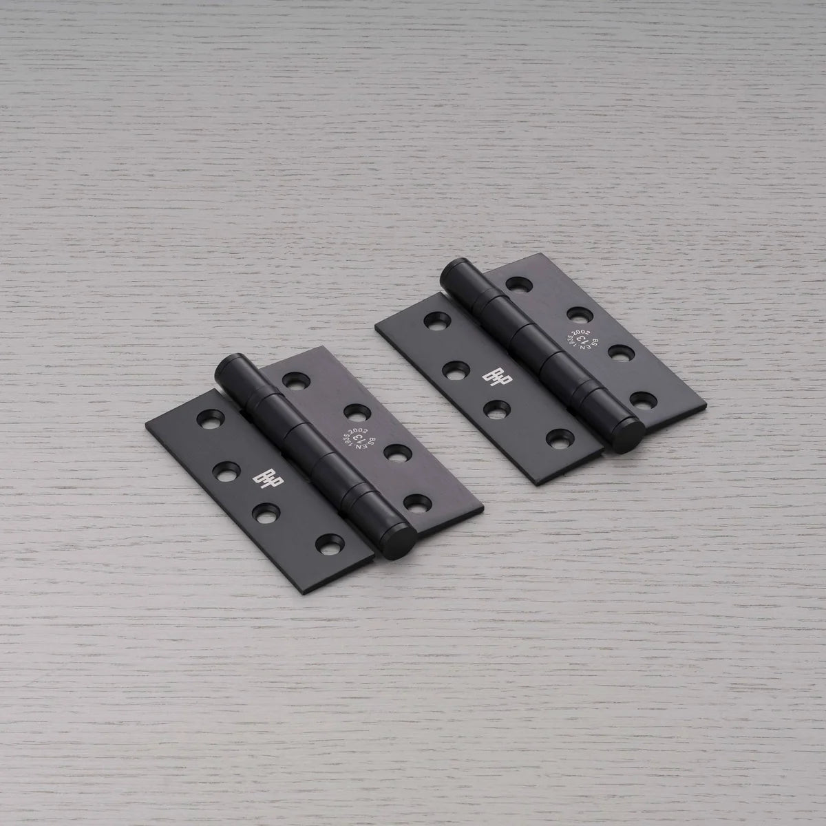 Pair of Buster + Punch black door hinges laid on a pale grey wood grain surface, with countersunk screw holes